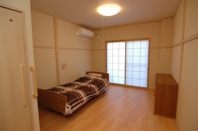 介護施設の居室。ベッド、木製家具、障子窓、明るい照明が整備された個室。